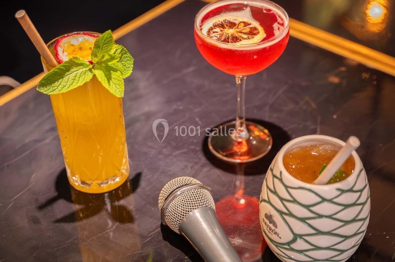 Trois cocktails colorés sur une table avec un micro en premier plan, ambiance de bar ou de soirée.