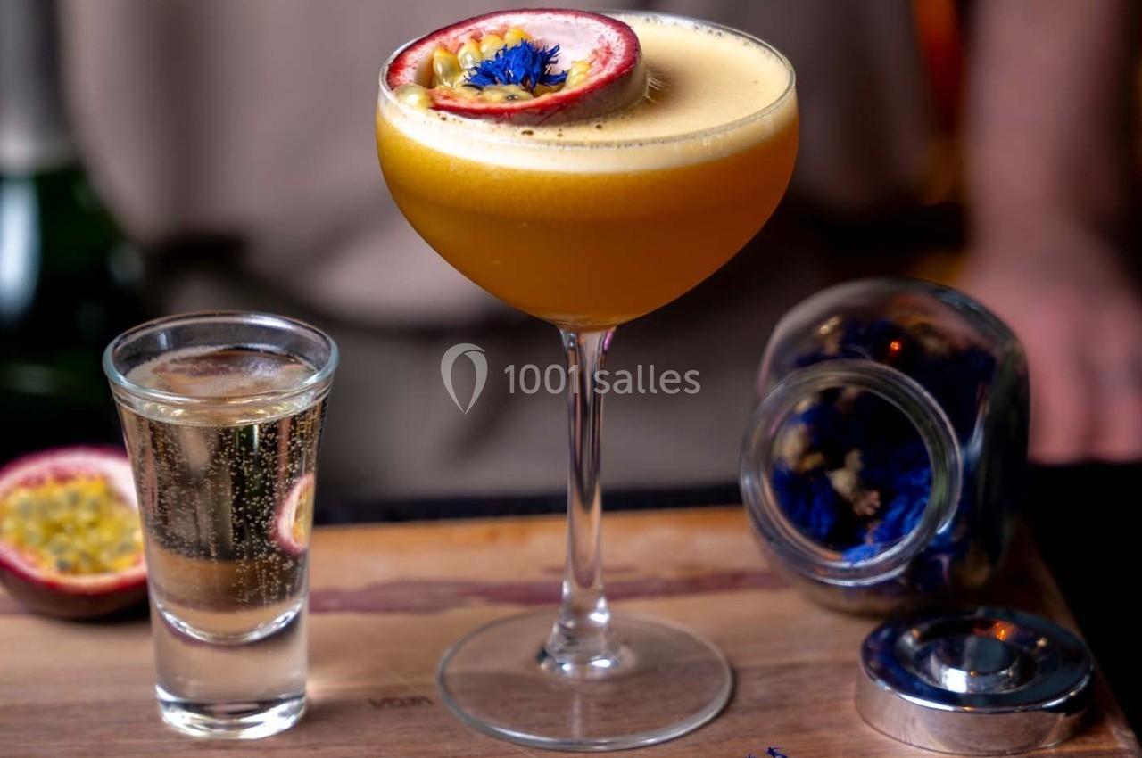 Cocktail jaune orangé garni d’un demi-fruit de la passion, accompagné d’un verre de champagne sur un comptoir en bois.