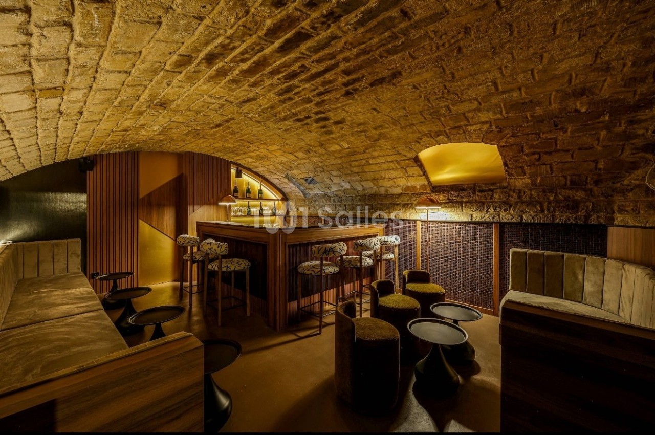 Salle voûtée en pierre avec un bar central en bois, tabourets et éclairage chaleureux.
