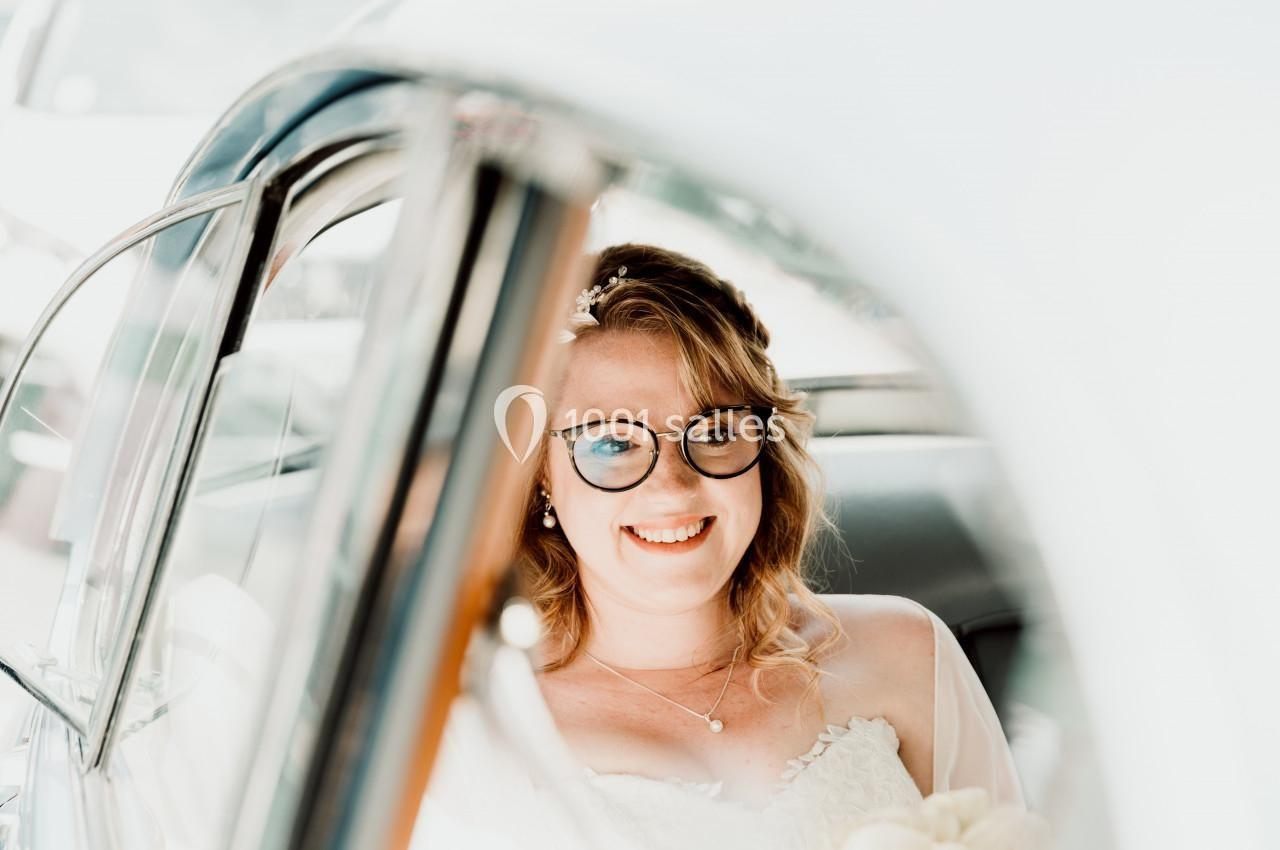Une femme souriante en robe blanche est assise à l'intérieur d'une voiture ancienne, vue à travers une vitre.
