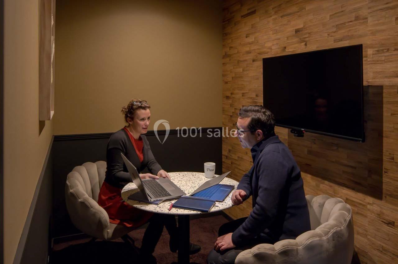 Deux personnes discutent autour d'une table ronde dans une petite salle avec un écran mural et des ordinateurs portables.