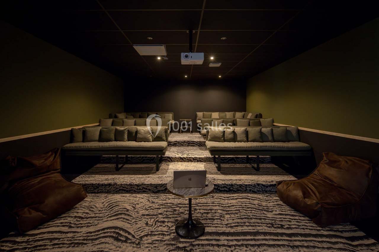 Salle de projection sombre avec plusieurs rangées de sièges rembourrés, poufs en cuir et un projecteur au plafond.