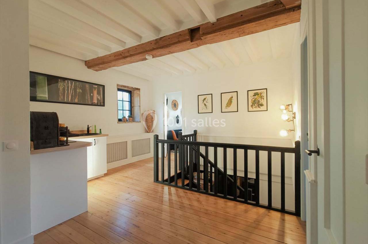 Vue d'un palier lumineux avec parquet en bois, poutres apparentes, escalier noir et décorations murales encadrées.