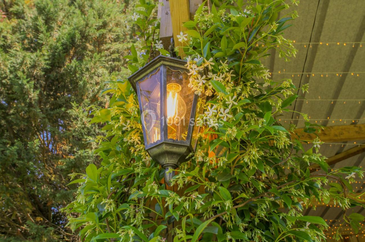 Lanterne allumée entourée de feuillage et de fleurs blanches, accrochée à une structure en bois sous un toit.