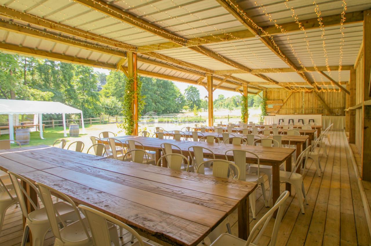 Salle de réception en bois ouverte sur l'extérieur, avec longues tables, chaises blanches et guirlandes lumineuses.