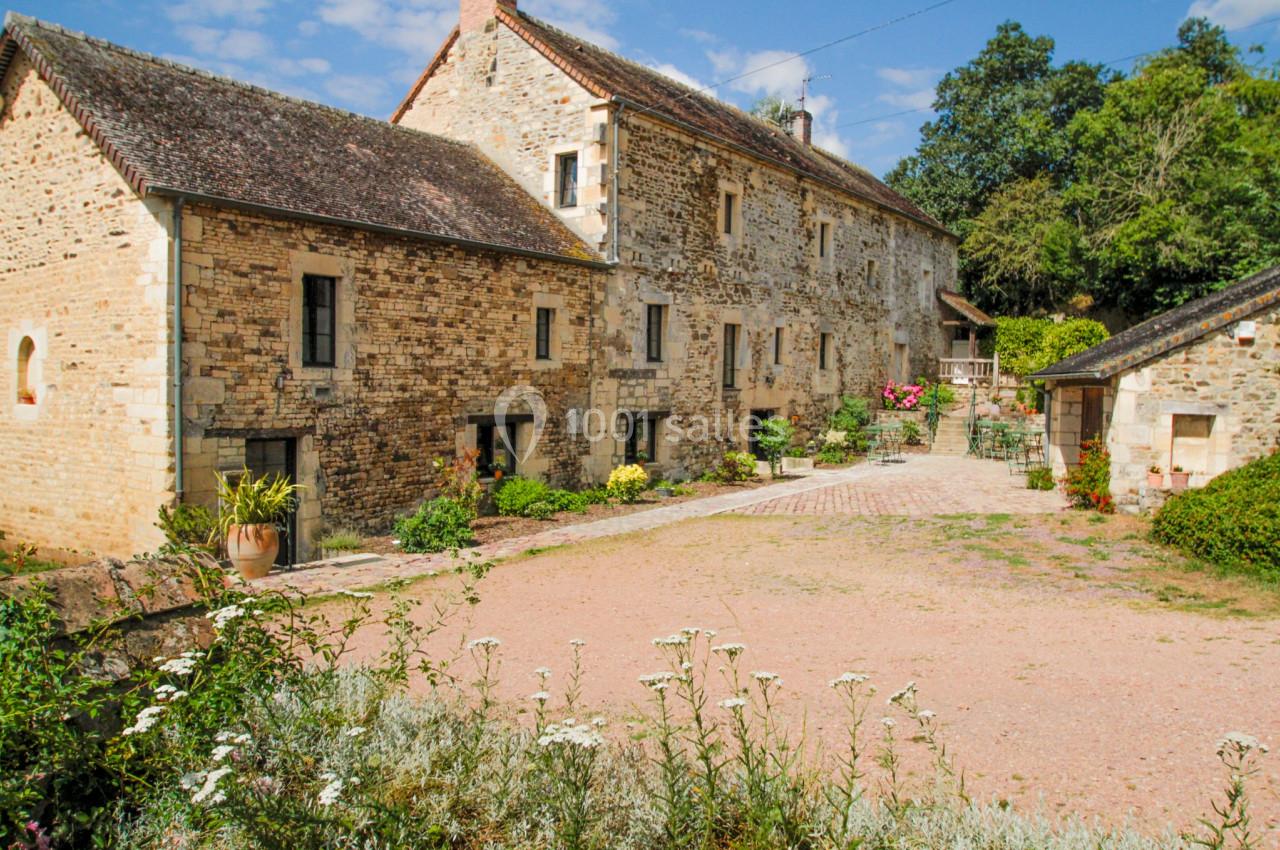 Cour pavée devant une maison en pierre ancienne avec des volets en bois, entourée de végétation et fleurs.
