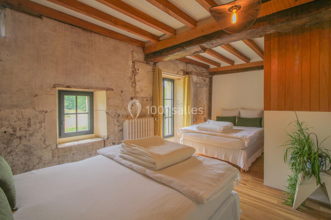 Chambre lumineuse avec deux lits doubles, murs en pierre apparente, poutres en bois et une plante décorative.