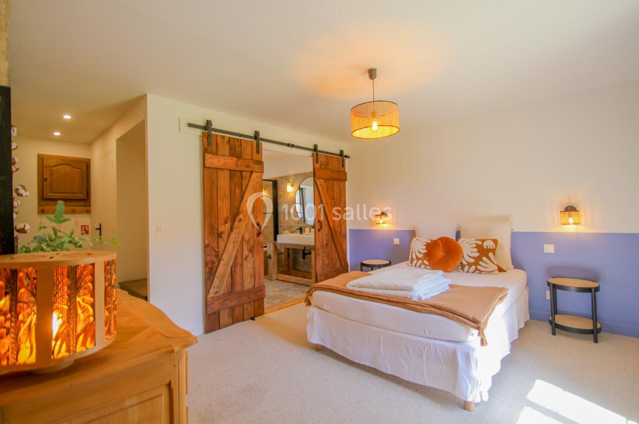 Chambre lumineuse avec lit double, portes coulissantes en bois, et salle de bain attenante visible en arrière-plan.