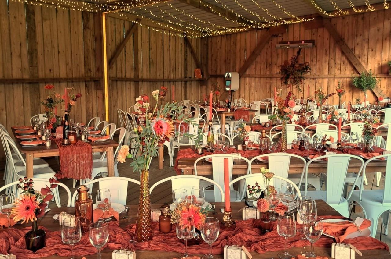 Salle décorée pour un événement, avec tables dressées, nappes rouges, fleurs orange et guirlandes lumineuses suspendues.