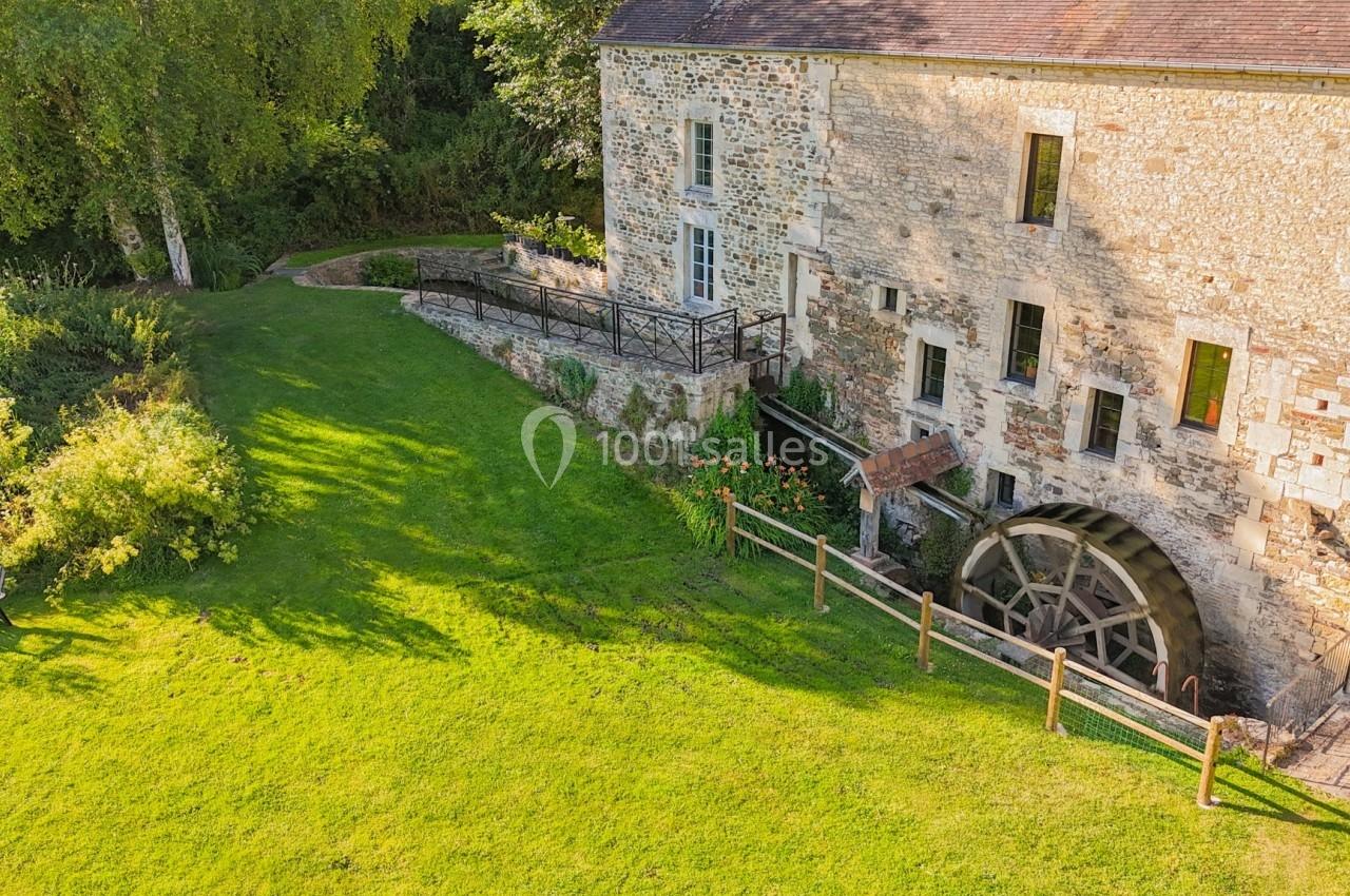 Vue d'un ancien moulin en pierre avec une roue à aubes, entouré d'un jardin verdoyant et d'arbres.