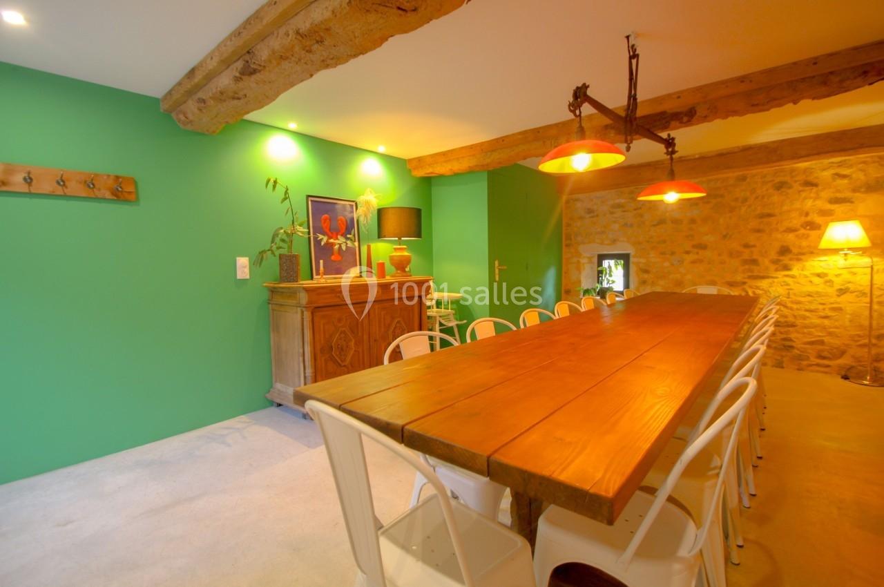 Salle à manger avec grande table en bois, chaises blanches, mur vert, poutres apparentes et éclairage chaleureux.