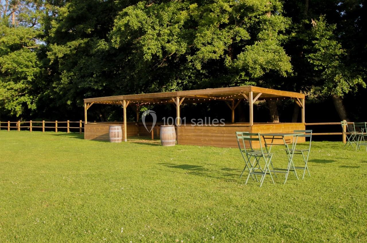 Abri en bois avec guirlandes lumineuses, entouré de tables et chaises sur une pelouse, bordé d'arbres et d'une clôture.