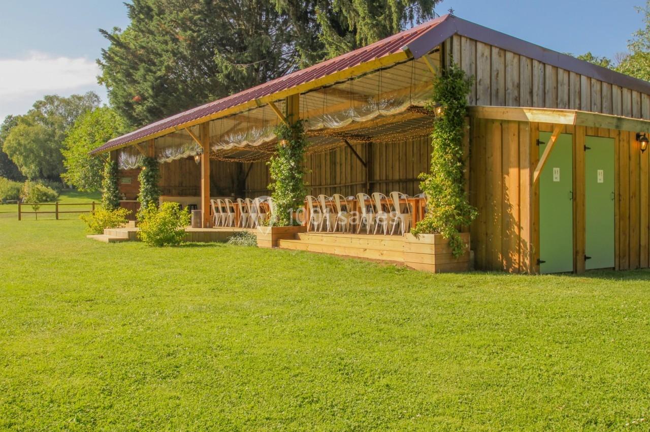 Abri en bois avec des chaises alignées sous une pergola, entouré de verdure et éclairé par la lumière du jour.