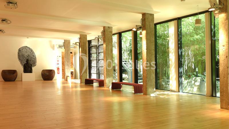 Salle lumineuse avec parquet en bois, grandes baies vitrées donnant sur un jardin, colonnes en béton et œuvres d'art…