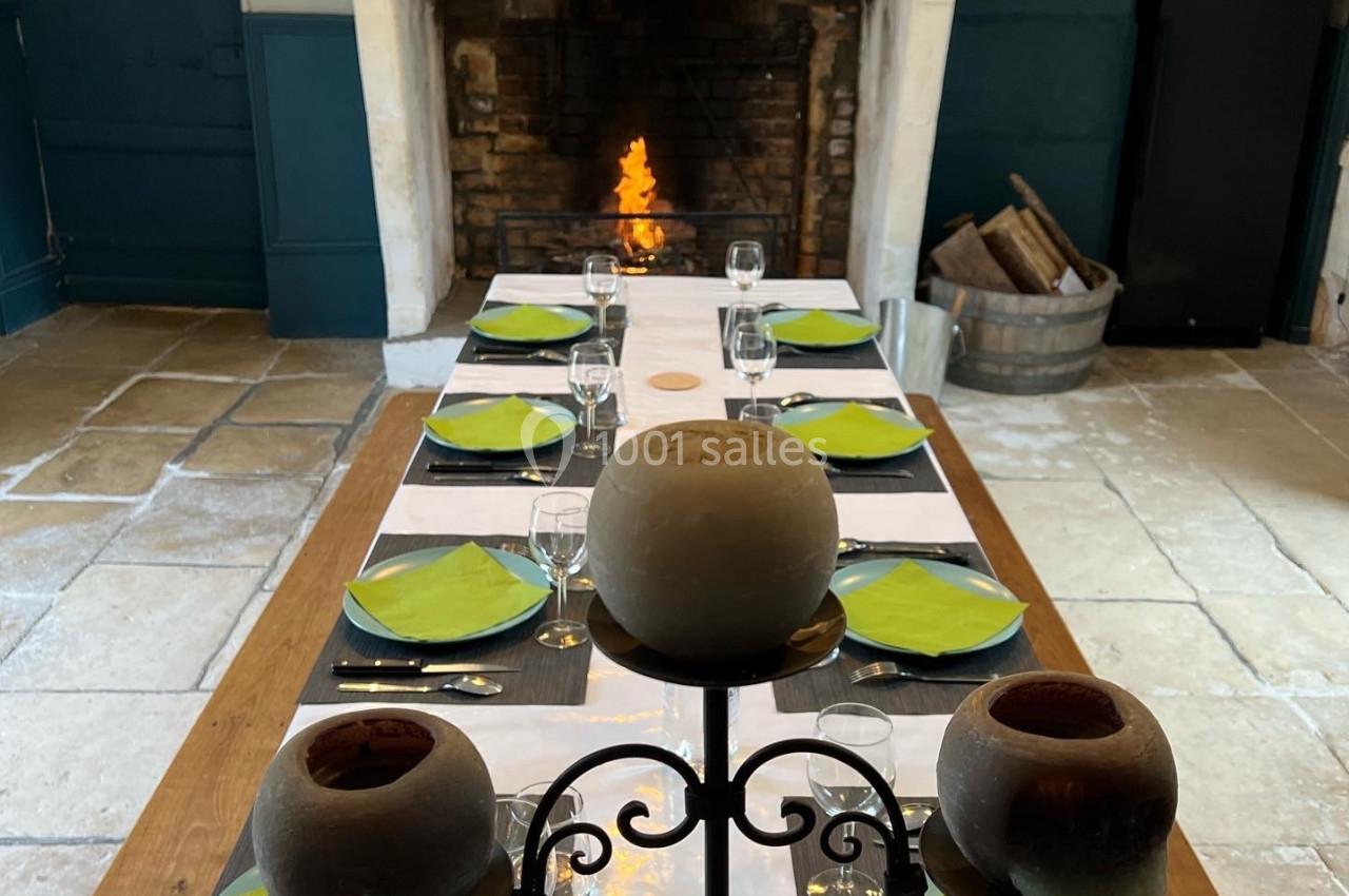 Table dressée avec des assiettes vertes et des verres, devant une cheminée allumée dans une pièce au sol en pierre.
