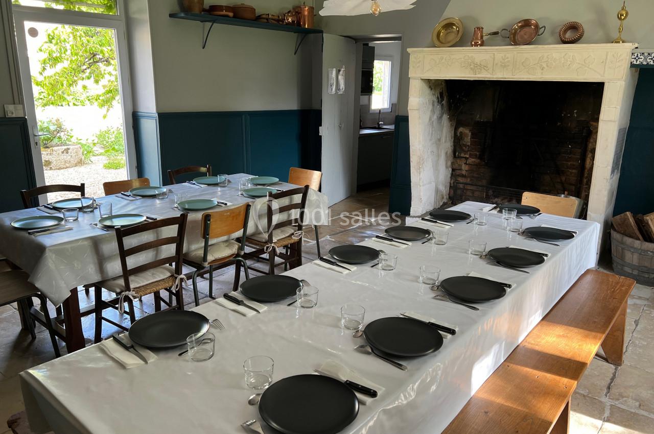 Grande table dressée avec des assiettes noires et des couverts dans une salle à manger rustique avec cheminée et lumière…