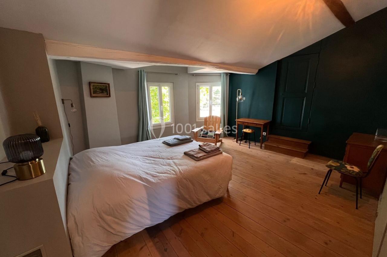 Chambre lumineuse avec lit double, parquet en bois, bureau, chaise et mur vert foncé, éclairée par une fenêtre.