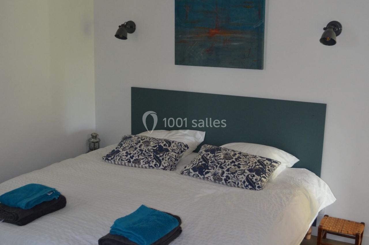 Lit double avec tête de lit verte, draps blancs, coussins à motifs, serviettes pliées et éclairage mural dans une chambre…