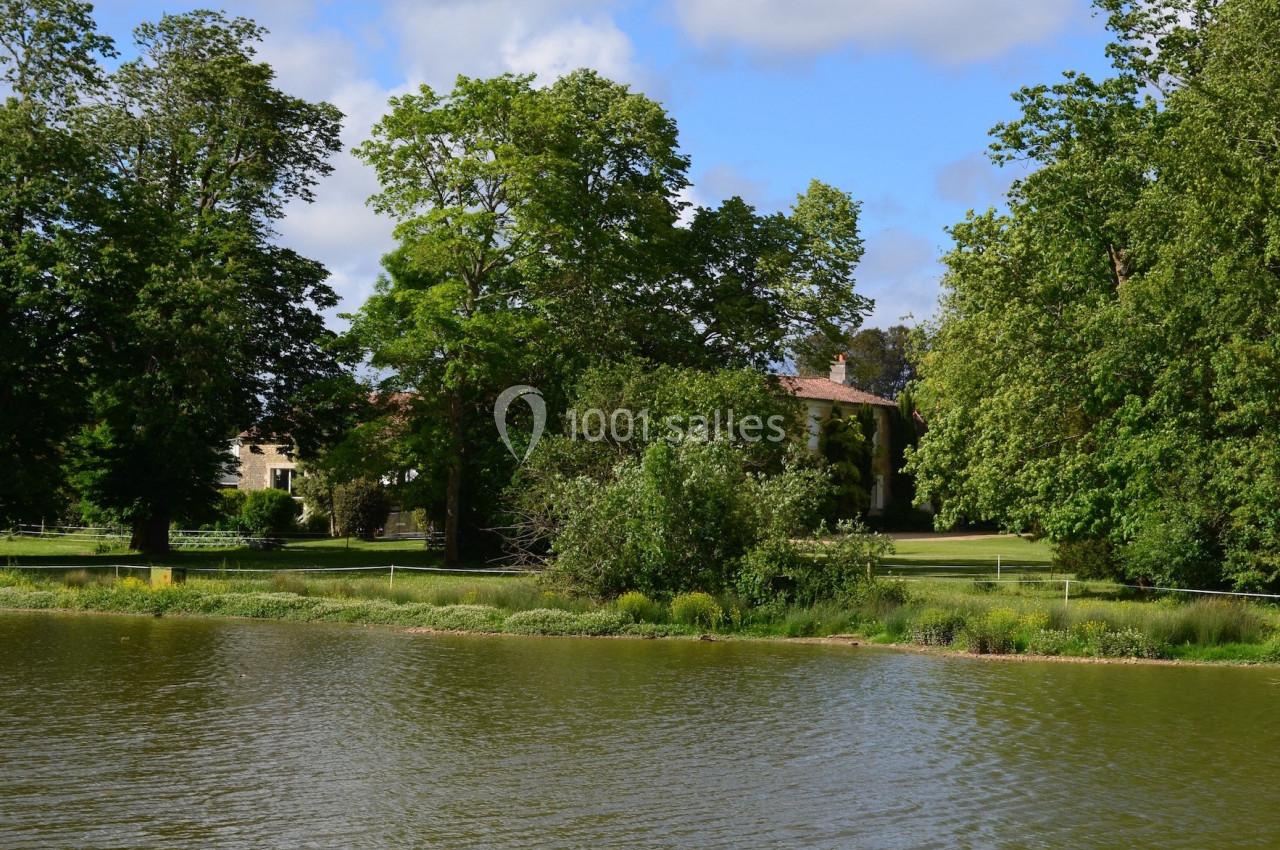 Vue d'un lac entouré de verdure avec des arbres et une maison en arrière-plan sous un ciel partiellement nuageux.