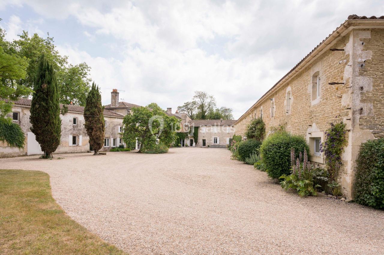 Cour intérieure d'un domaine en pierre avec bâtiments anciens, arbres et allée gravillonnée sous un ciel partiellement…