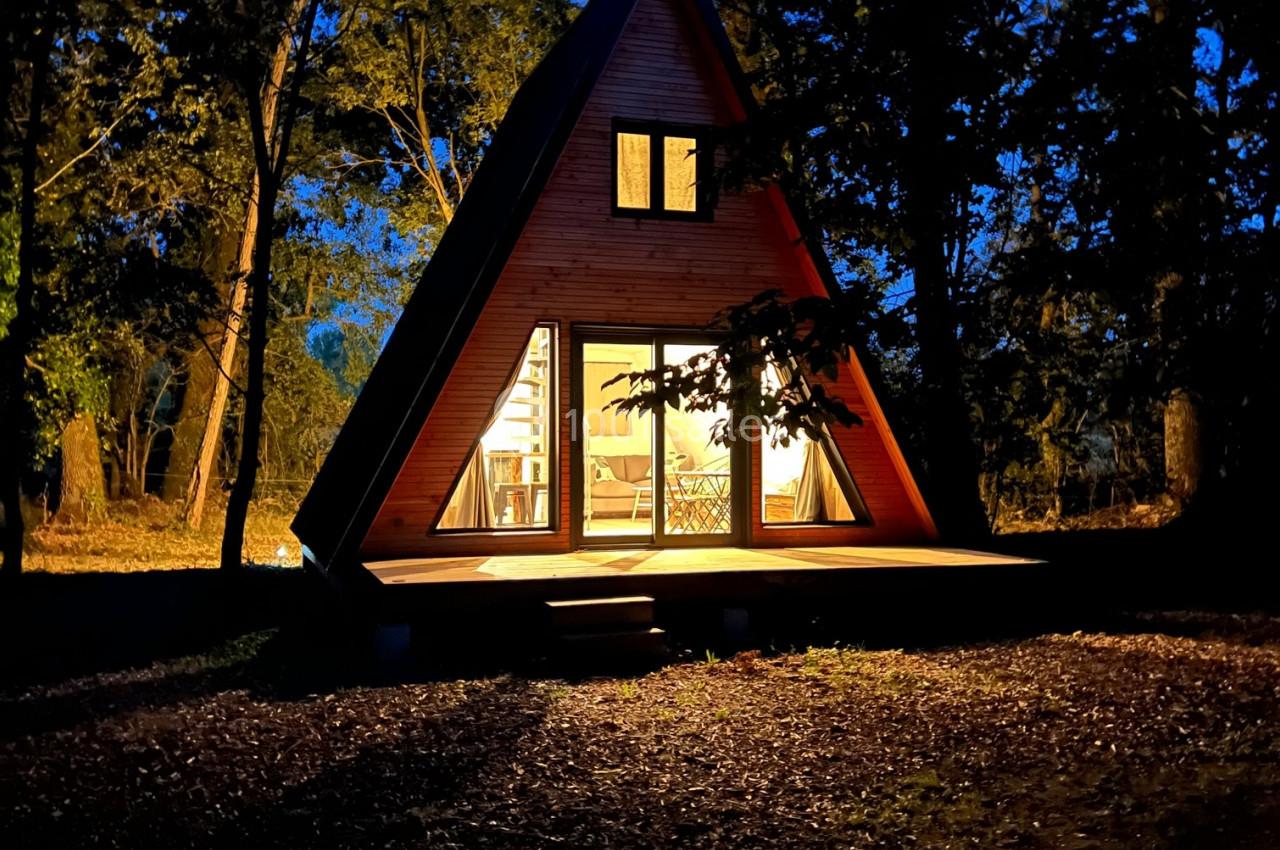 Cabane en forme de A éclairée de l'intérieur, entourée d'arbres dans un environnement nocturne.