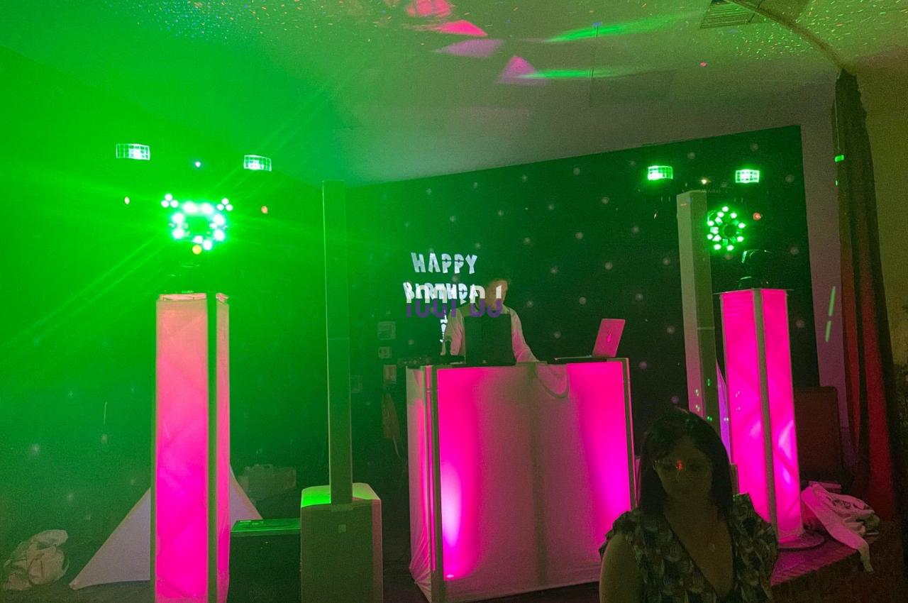Un DJ anime une soirée avec des lumières vertes et roses, une scène éclairée et un panneau ’Happy Birthday’.