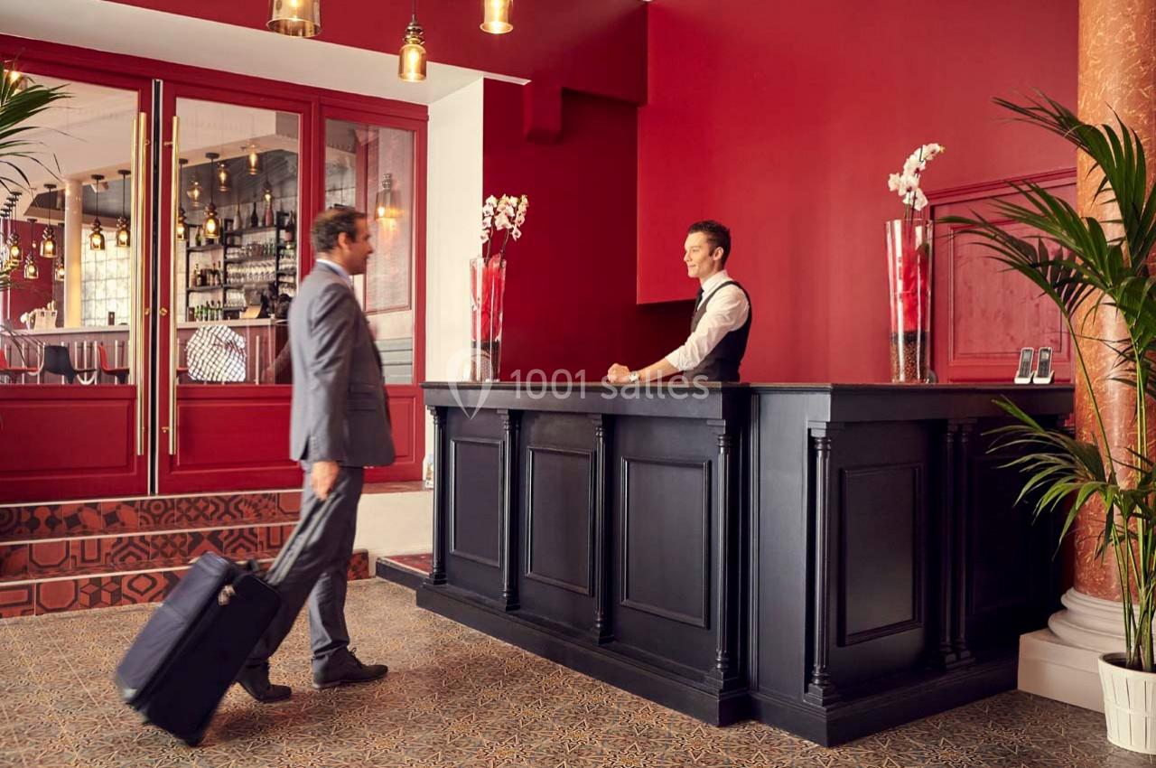 Un homme en costume avec une valise s'approche du comptoir de réception d'un hôtel au décor élégant et chaleureux.