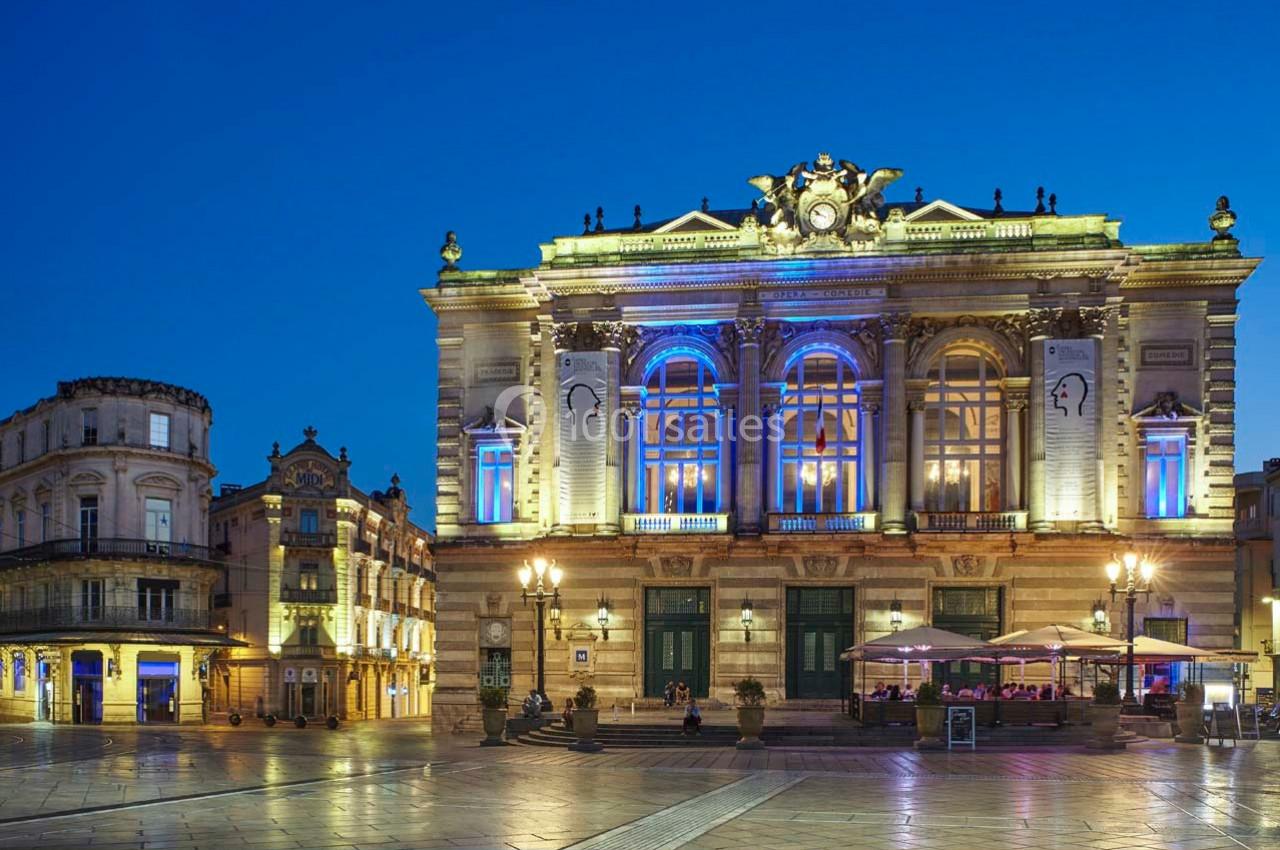 Façade illuminée d'un théâtre historique avec terrasse de café en soirée sur une place pavée.
