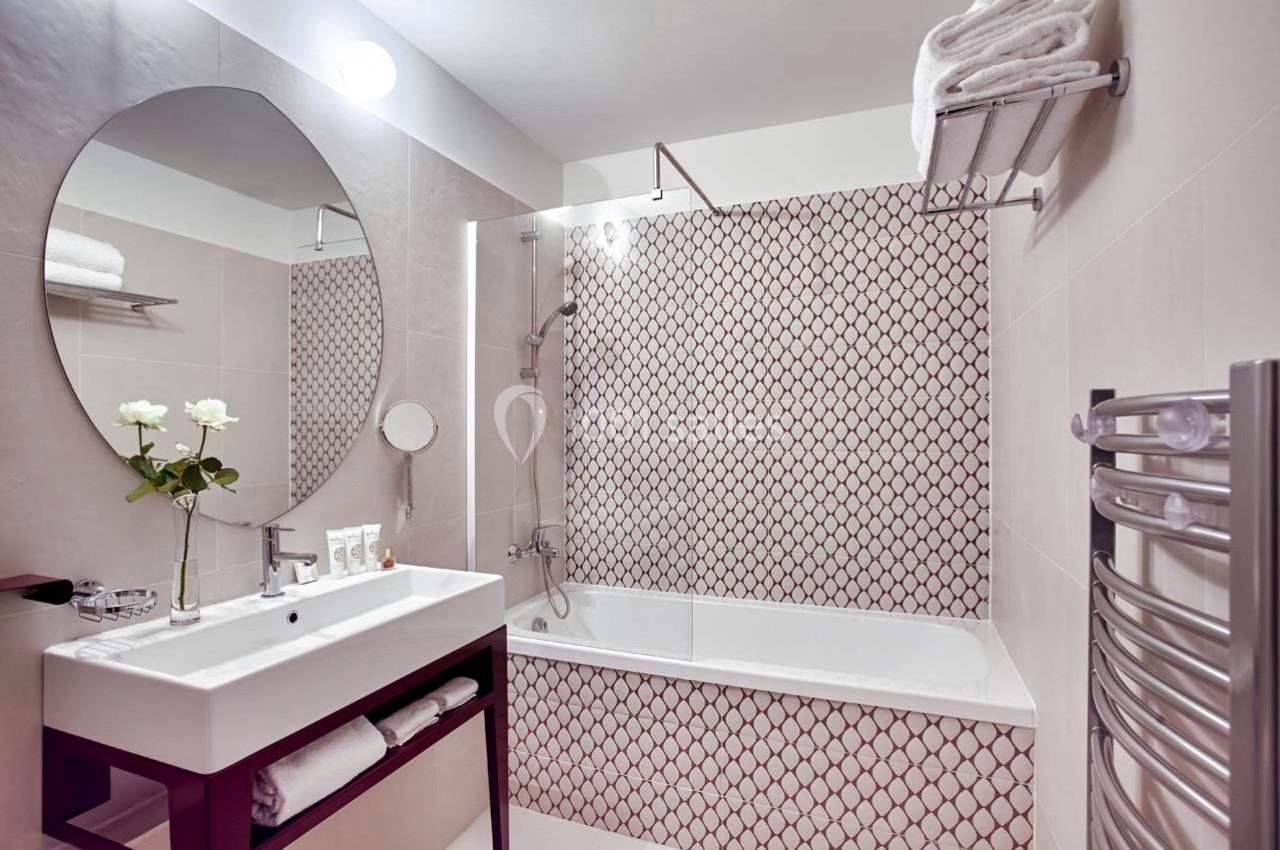 Salle de bain avec baignoire, carrelage à motifs, lavabo avec miroir ovale, et porte-serviettes mural.