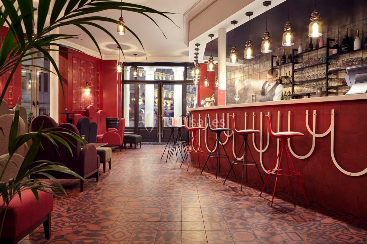 Intérieur d'un bar avec comptoir rouge, tabourets hauts, éclairage suspendu et fauteuils dans un décor chaleureux.