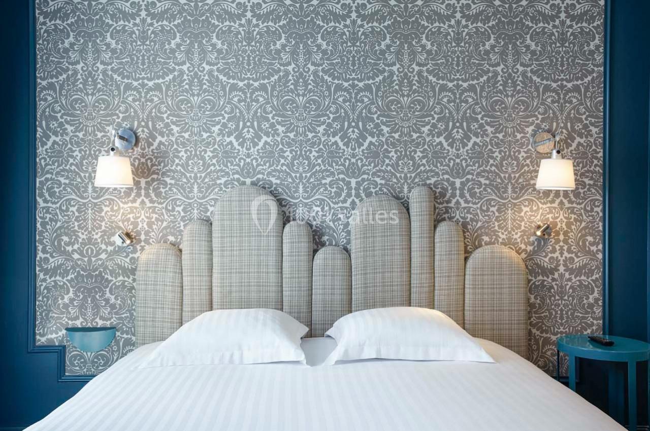 Lit double avec tête de lit capitonnée à motifs géométriques, encadré par deux appliques murales et un mur à motifs floraux.