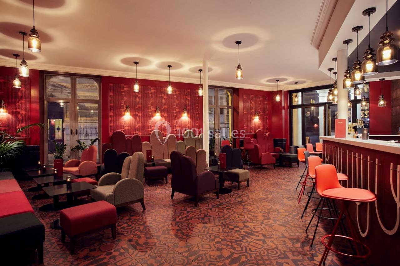 Salle lumineuse avec fauteuils colorés, tables basses, bar moderne et décor rouge chaleureux.