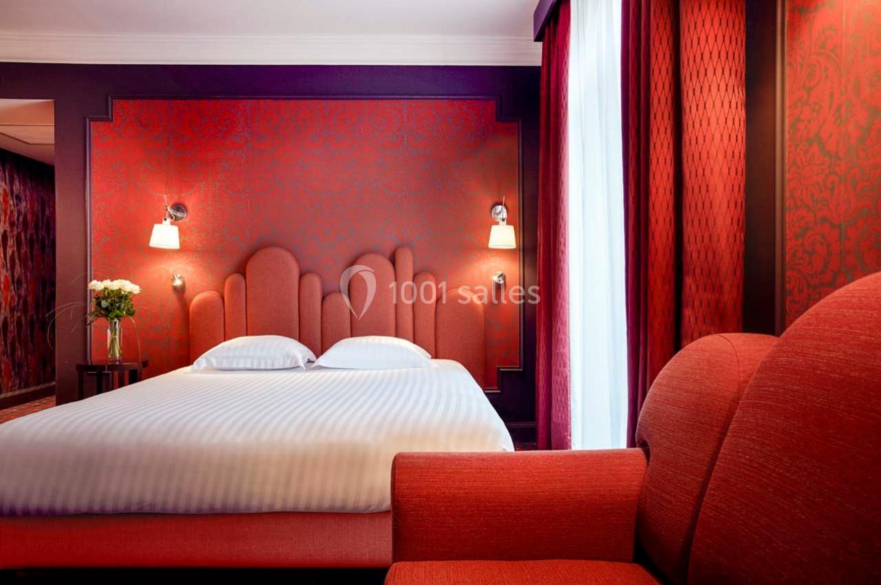 Chambre d'hôtel avec lit double, tête de lit rouge, éclairage mural, fauteuil assorti et rideaux rouges.