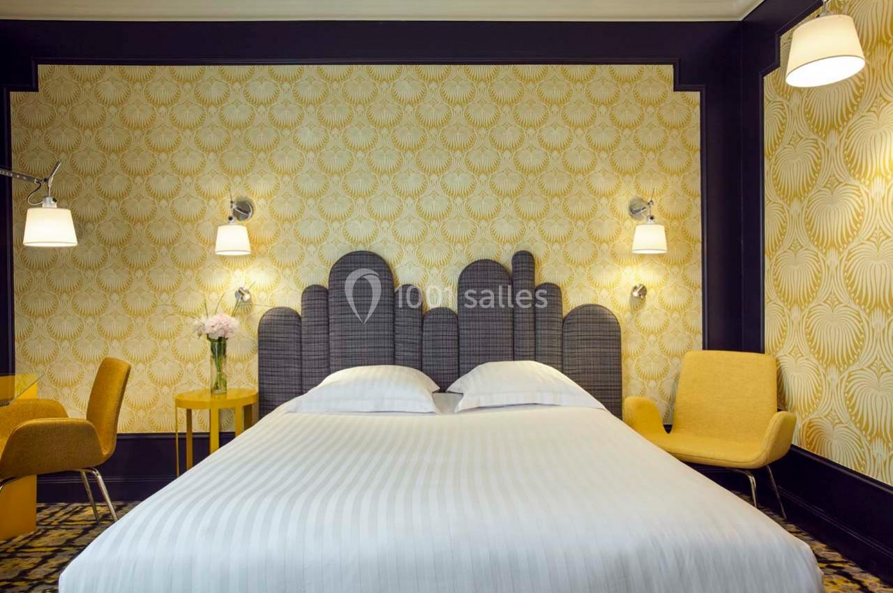 Chambre lumineuse avec lit double, tête de lit grise, fauteuils jaunes, lampes murales et papier peint à motifs dorés.