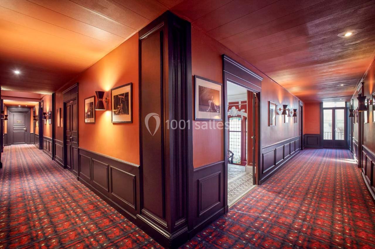 Couloir d'hôtel avec moquette rouge, murs orange, éclairage mural et portes menant à différentes pièces.