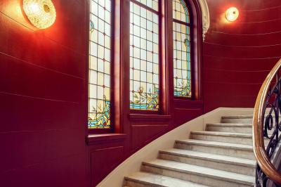 Escalier en marbre avec une rampe en bois, éclairé par des vitraux colorés et des appliques murales sur des murs rouges.