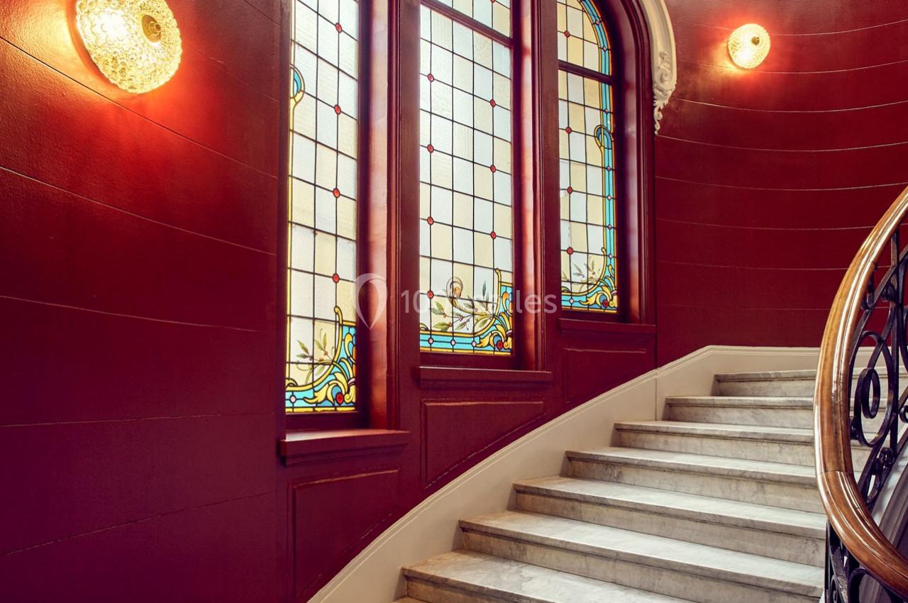 Escalier en marbre avec une rampe en bois, éclairé par des vitraux colorés et des appliques murales sur des murs rouges.