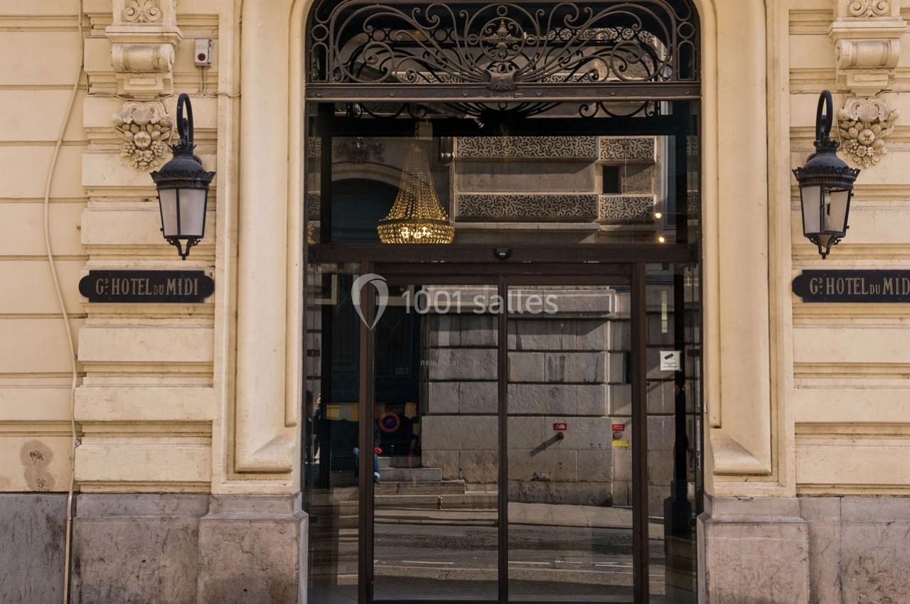 Entrée d'un hôtel avec une porte vitrée, des lampes murales et un lustre visible à l'intérieur.