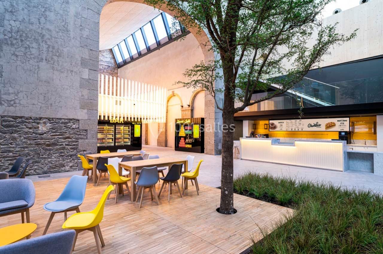 Espace moderne avec tables, chaises colorées, arbre central, murs en pierre et comptoir lumineux en arrière-plan.