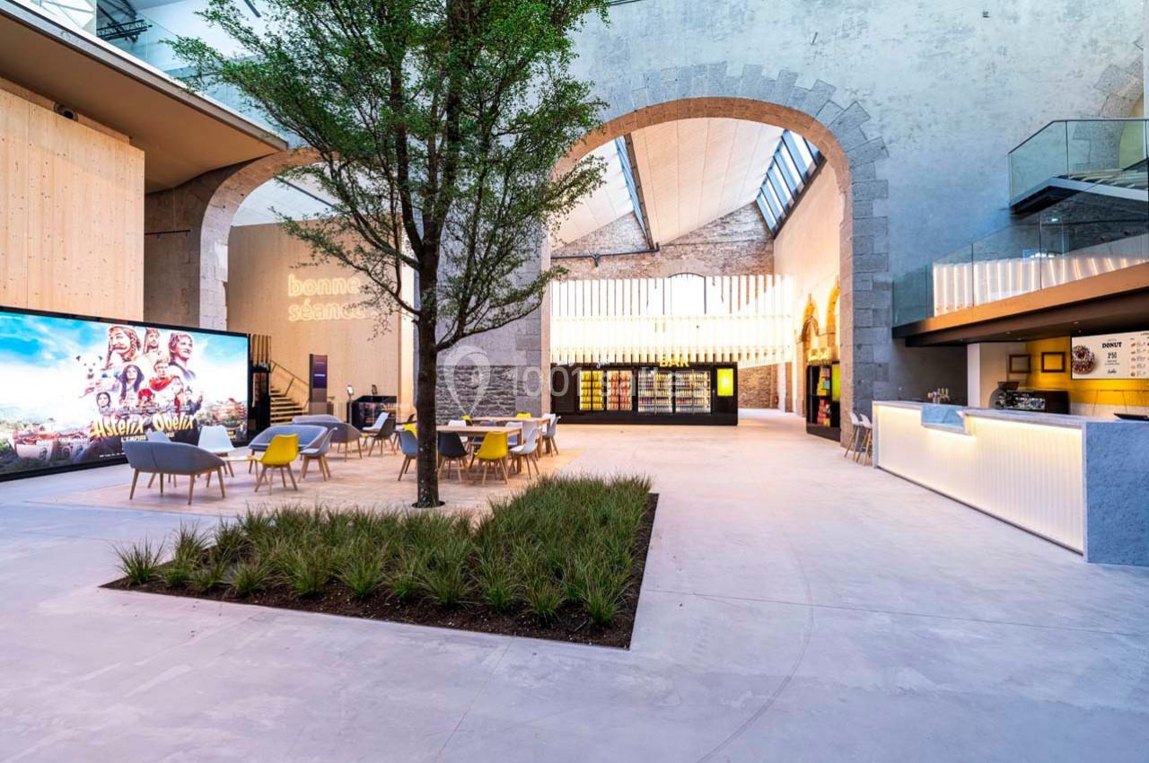 Espace intérieur moderne avec un arbre central, des sièges, un comptoir lumineux et un écran affichant une image.