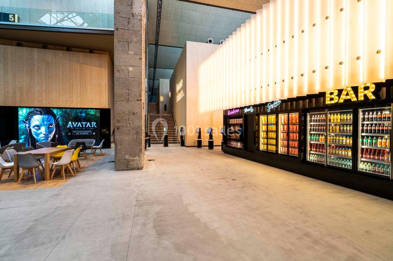 Espace moderne avec distributeurs de boissons, tables, chaises et affichage numérique dans un hall lumineux.