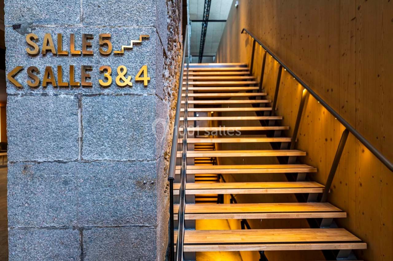 Escalier en bois éclairé menant aux salles 3, 4 et 5, avec des panneaux directionnels sur un mur en pierre.