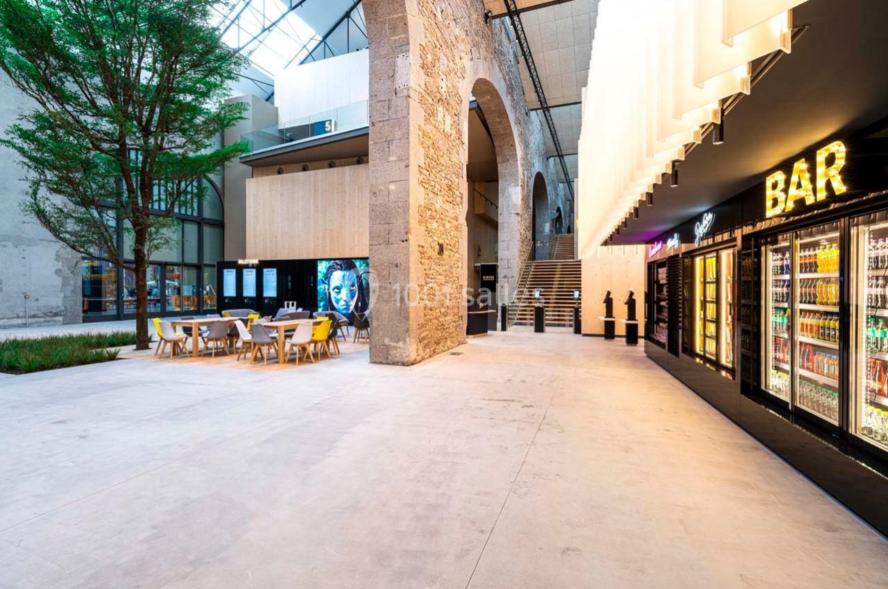 Espace intérieur moderne avec tables, arbre, arc en pierre, distributeurs de boissons et enseigne lumineuse ’BAR’.