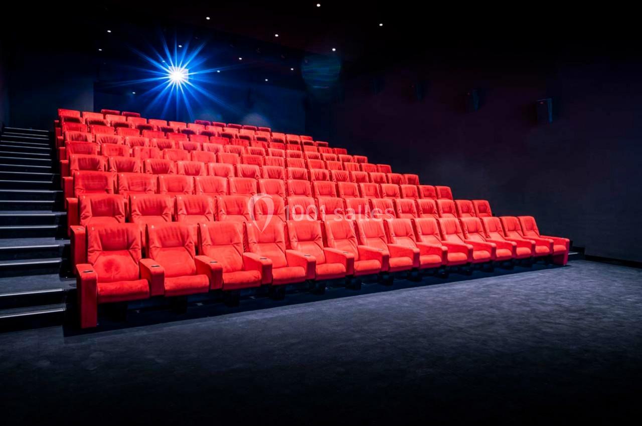 Salle de cinéma vide avec des rangées de fauteuils rouges et un projecteur allumé au fond.