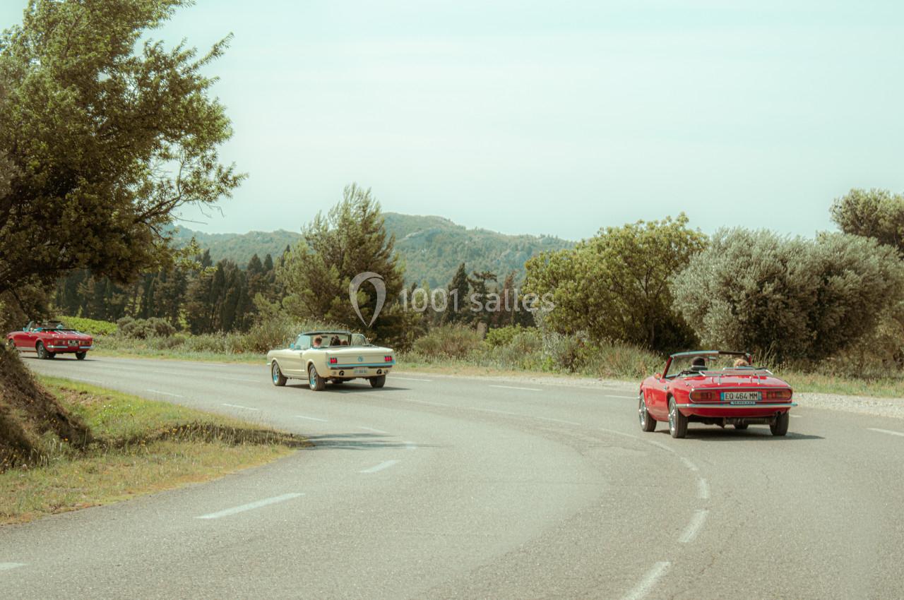 Trois voitures décapotables anciennes circulent sur une route sinueuse entourée de végétation et de collines.