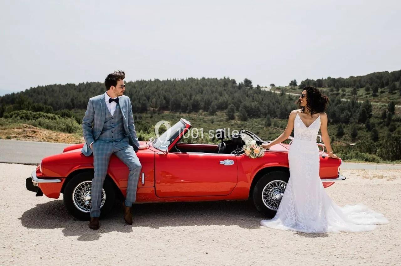 Un couple en tenue de mariage pose près d'une voiture décapotable rouge dans un paysage naturel.