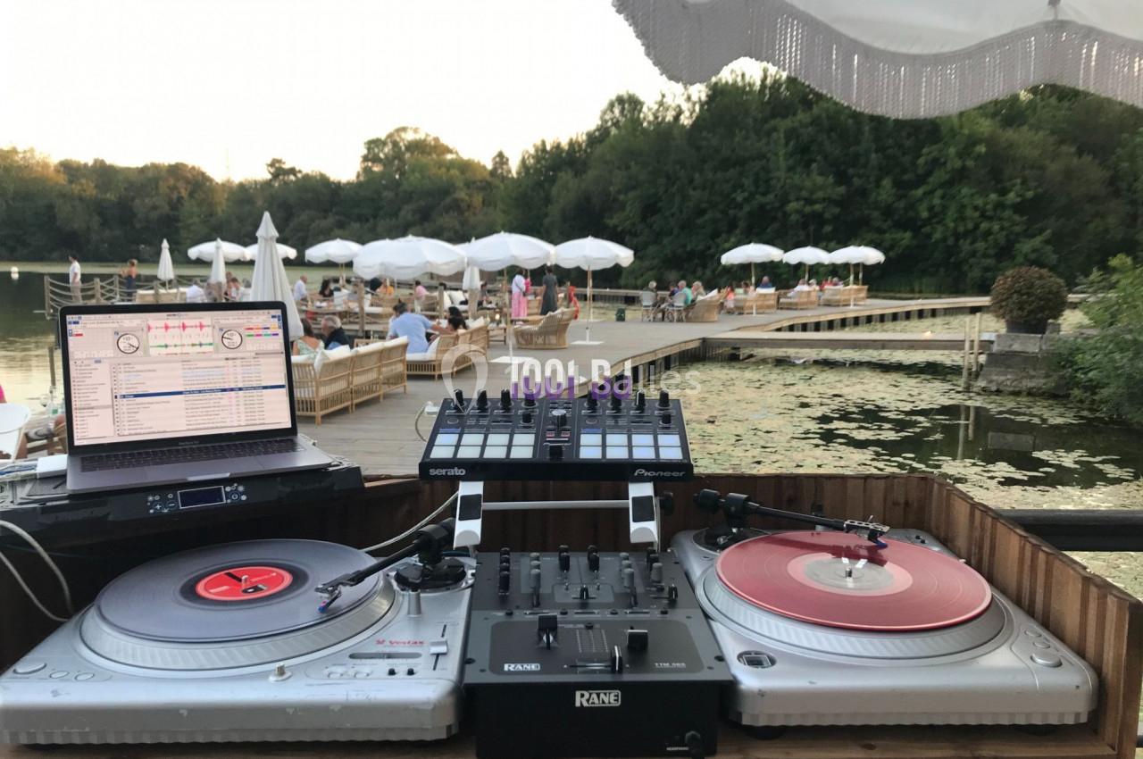 Poste de DJ en plein air avec platines, table de mixage et ordinateur, face à un lac entouré de végétation et de parasols.