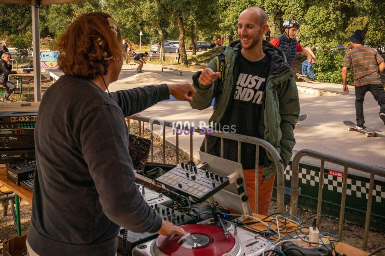 Un DJ serre le poing d'un homme souriant près d'une table de mixage, avec des skateurs en arrière-plan.