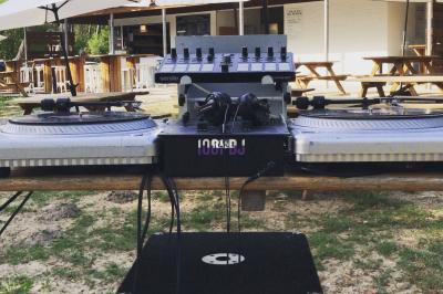 Deux platines vinyles et une table de mixage installées en extérieur sous des parasols, avec des tables en arrière-plan.