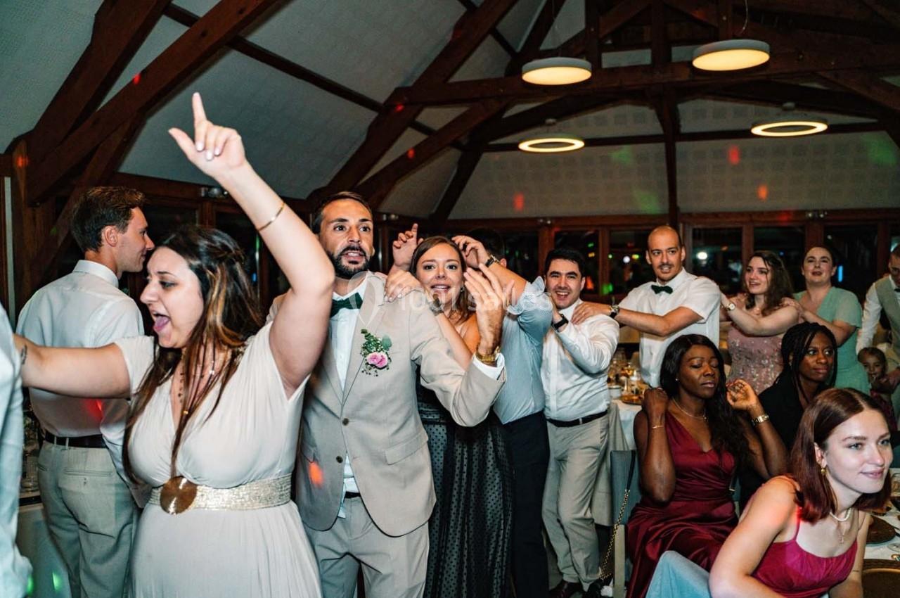 Des invités dansent et célèbrent dans une salle avec charpente en bois, éclairée par des suspensions circulaires.